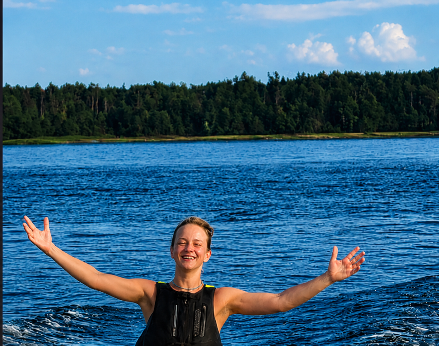 Naerev naine mustas päästevestis käed laiali sirutatud, taga sinine merevesi, metsane rannajoon ja pilvine suvetaevas